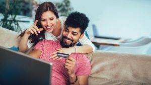 young adult couple sitting together on couch shopping online with laptop and debit card