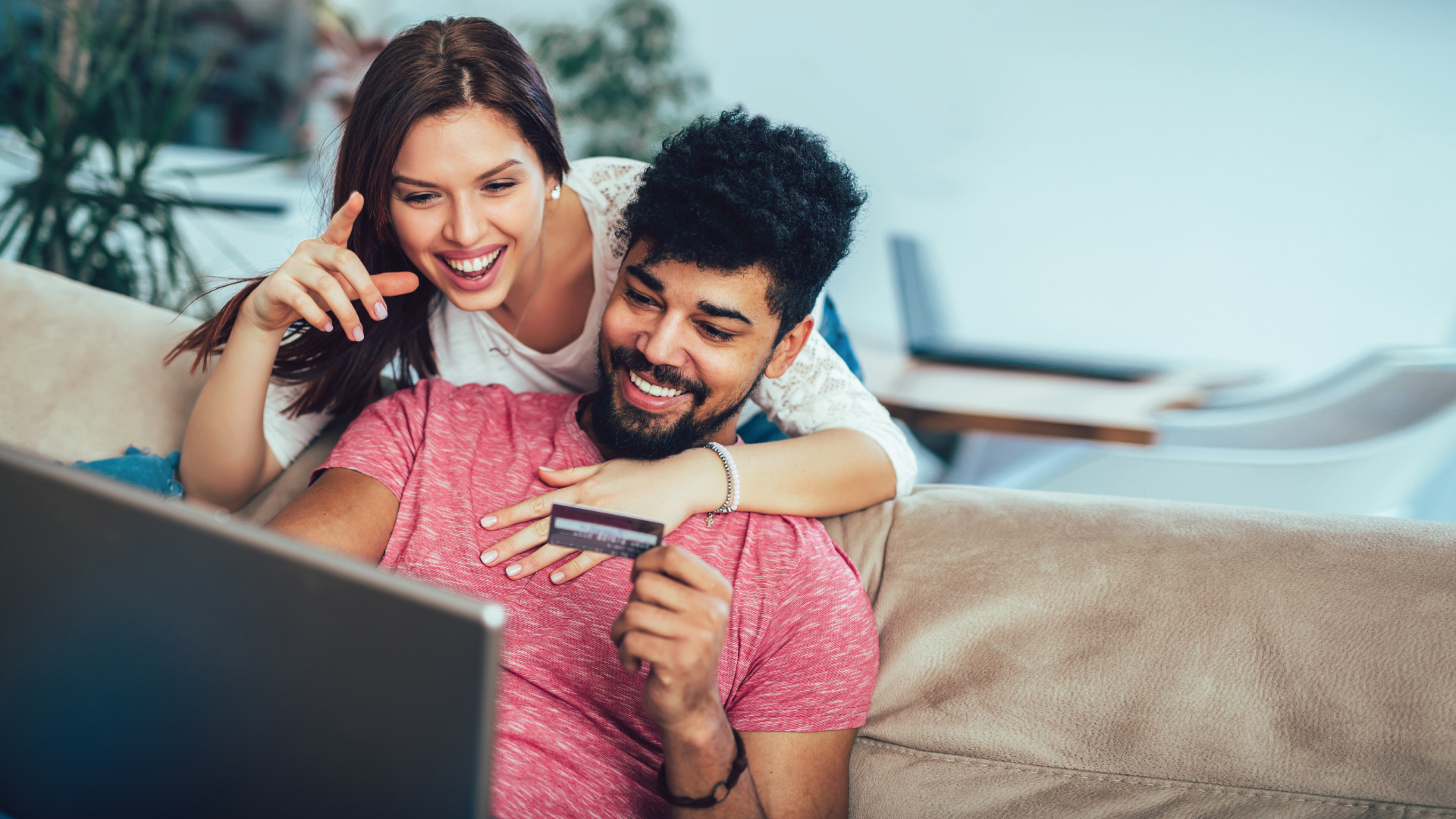 young adult couple sitting together on couch shopping online with laptop and debit card