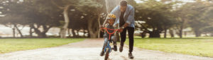 Father teaching young son to ride bicycle, young son on red bicycle wearing helmet, both smiling brightly