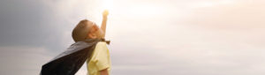 Young child with cape and goggles with arm raised in superhero pose