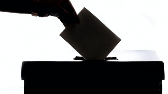 Person dropping a piece of paper into a voting ballot box.